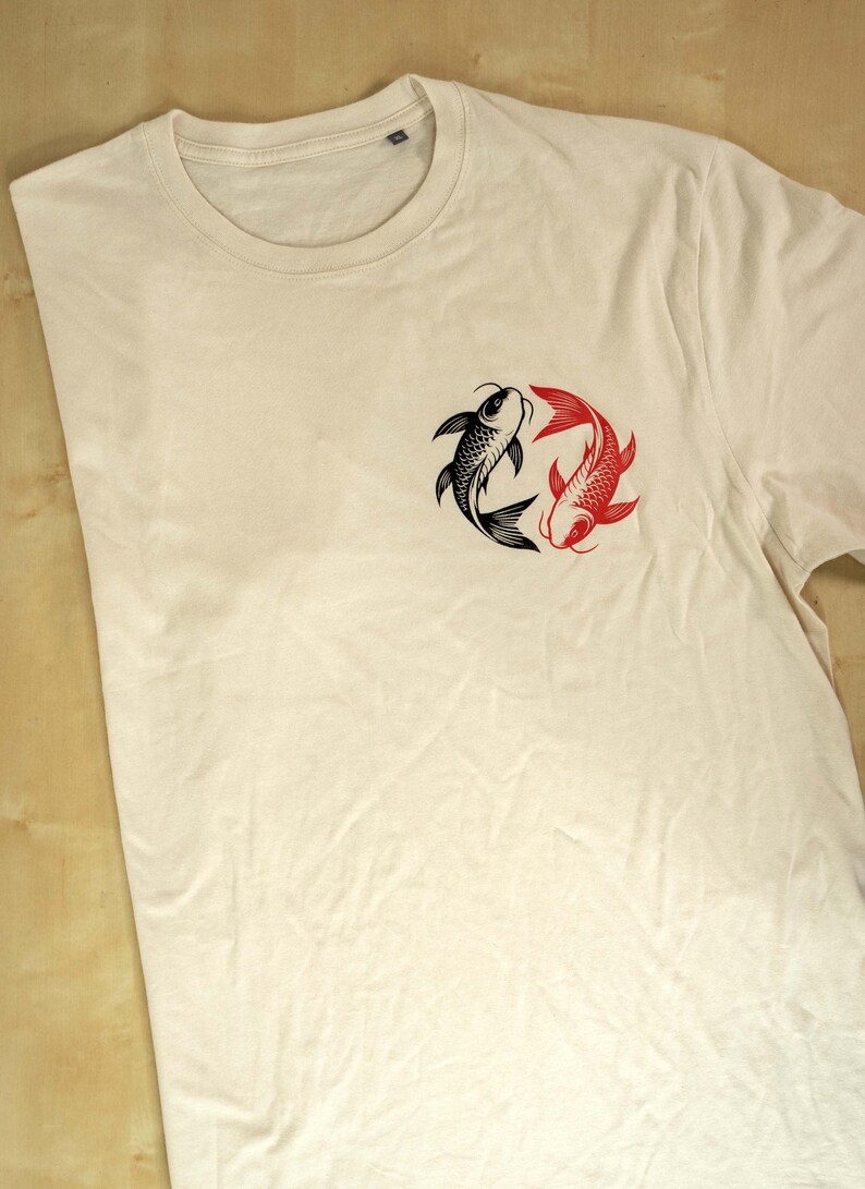 K&ouml;nnte beinhalten: Cremefarbenes T-Shirt mit einem schwarzen und roten Koi-Fisch-Design. Die Fische sind in einem kreisf&ouml;rmigen Muster angeordnet, wobei sich ein schwarzer und ein roter Fisch gegen&uuml;berstehen. Das Shirt hat einen Rundhalsausschnitt.