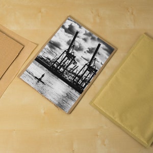 May include: A black and white photograph of a harbor scene with large cranes and a single person standing in the water. The photo is printed on a textured paper and is ready to be framed.