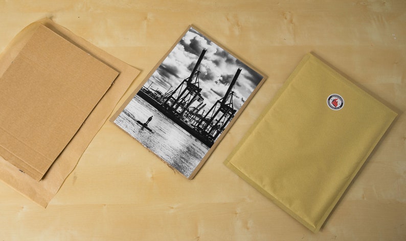 May include: A black and white photograph of a harbor scene with large cranes and a single person standing in the water. The photo is printed on a textured paper and is ready to be framed.