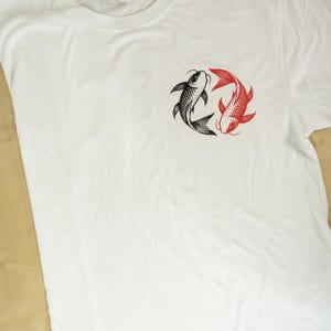 K&ouml;nnte beinhalten: Wei&szlig;es T-Shirt mit einem kreisf&ouml;rmigen Design von zwei Koi-Karpfen, einem schwarzen und einem roten. Die Fische sind im Yin-Yang-Stil angeordnet. Das T-Shirt besteht aus einem weichen Material, wahrscheinlich Baumwolle.