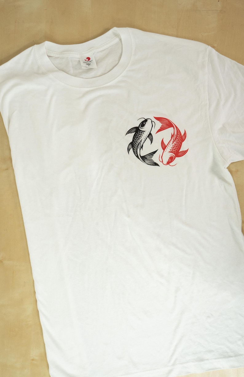 K&ouml;nnte beinhalten: Wei&szlig;es T-Shirt mit einem kreisf&ouml;rmigen Design von zwei Koi-Karpfen, einem schwarzen und einem roten. Die Fische sind im Yin-Yang-Stil angeordnet. Das T-Shirt besteht aus einem weichen Material, wahrscheinlich Baumwolle.