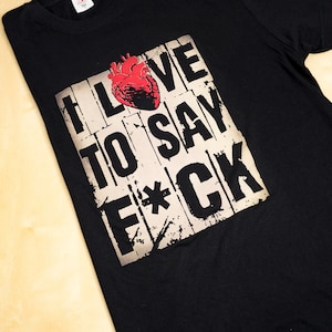 Könnte beinhalten: Schwarzes T-Shirt mit einem verwitterten Grafikdesign. Das Design zeigt ein rotes anatomisches Herz über dem Text "I LOVE TO SAY F*CK" in einer kräftigen, blockartigen Schrift. Das Shirt ist aus weichem Material.