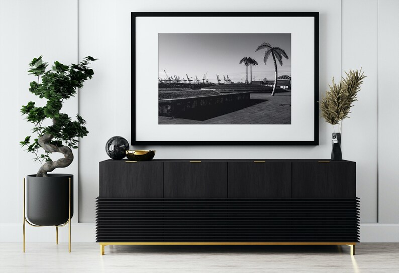 May include: A black and white photograph of a harbor with palm trees in the foreground, framed in a black frame and hanging above a black cabinet with gold accents.
