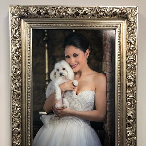 May include: A woman in a white wedding dress holds a small white dog in her arms. The woman is smiling and looking at the camera. The dog is looking at the woman. The photo is framed in a silver ornate frame.