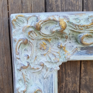 May include: A close-up of a decorative picture frame with a distressed, whitewashed finish and gold accents. The frame has a detailed, ornate design with a floral motif.