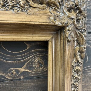 May include: A close-up of a decorative, ornate, wooden picture frame with a gold finish. The frame has a detailed, carved design with a floral motif.
