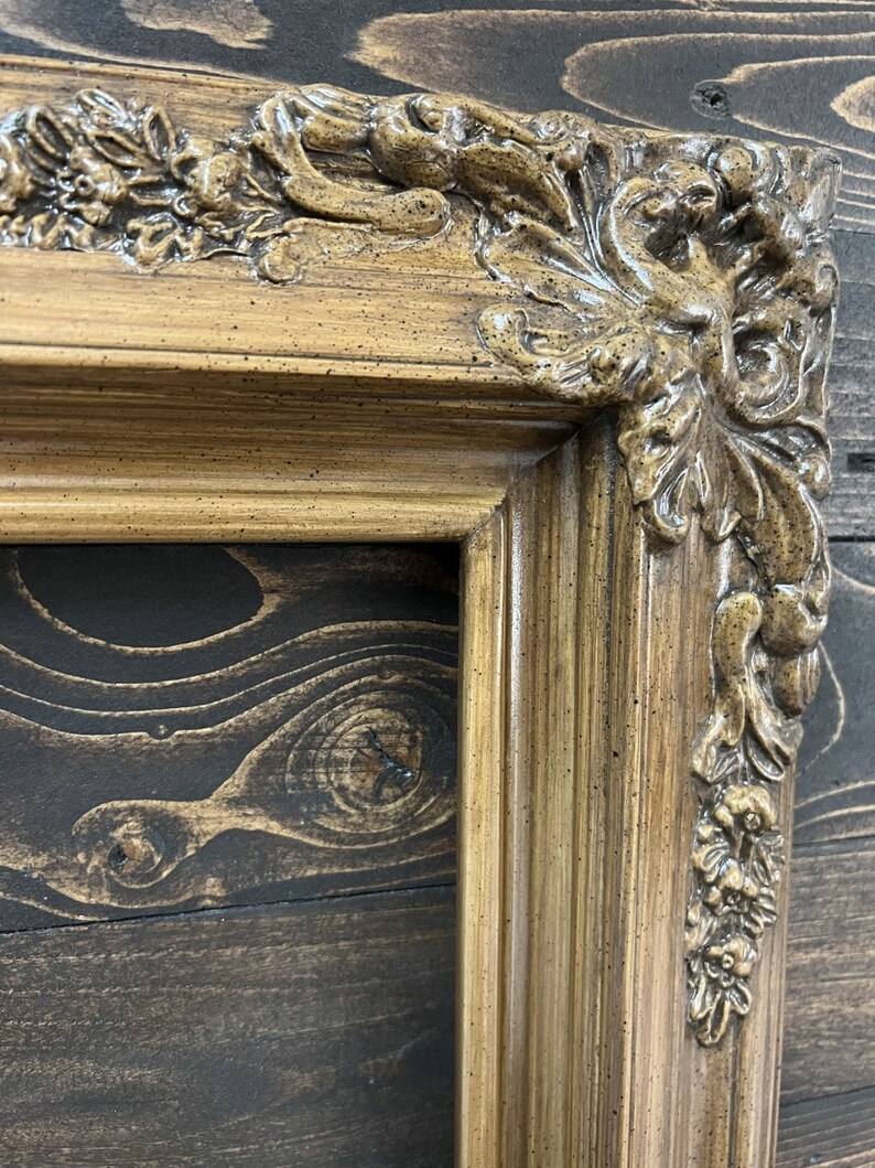 May include: A close-up of a decorative, ornate, wooden picture frame with a gold finish. The frame has a detailed, carved design with a floral motif.