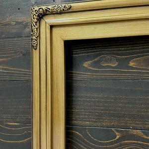 May include: A rectangular picture frame with a metallic gold finish and a dark, antiqued patina. The frame features a stepped profile and an ornate, raised floral scrollwork detail at the corner, showcasing a classic, multi-layered design.
