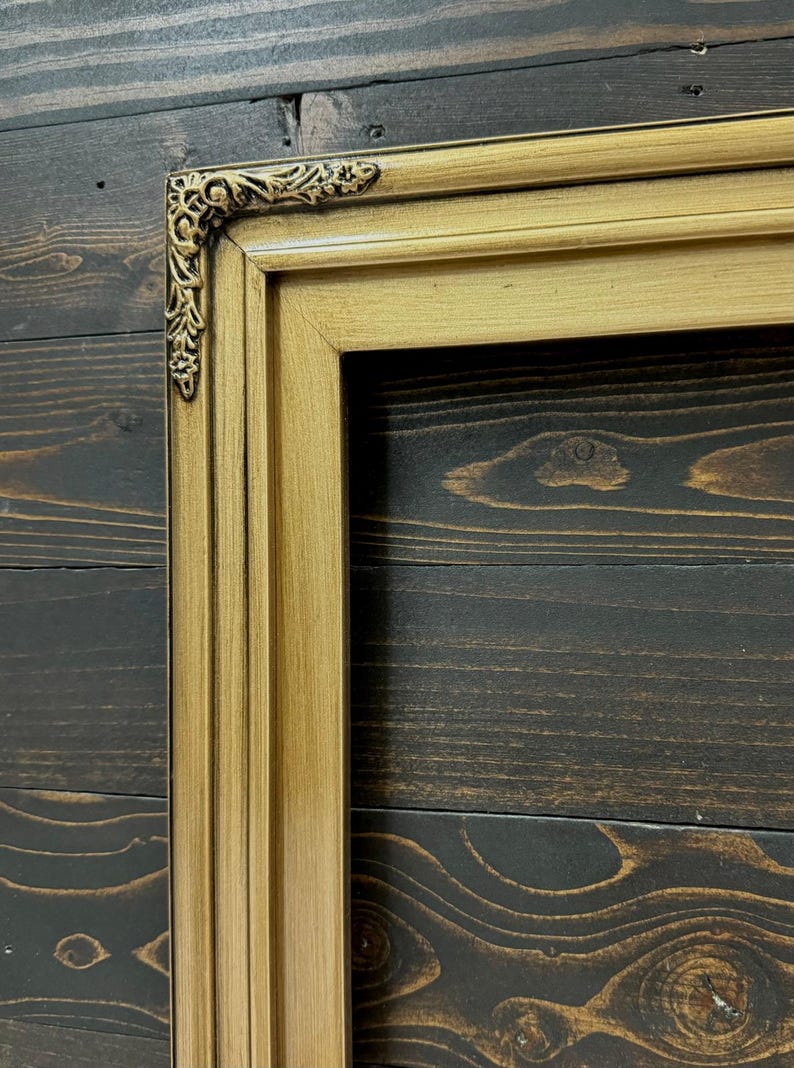 May include: A rectangular picture frame with a metallic gold finish and a dark, antiqued patina. The frame features a stepped profile and an ornate, raised floral scrollwork detail at the corner, showcasing a classic, multi-layered design.
