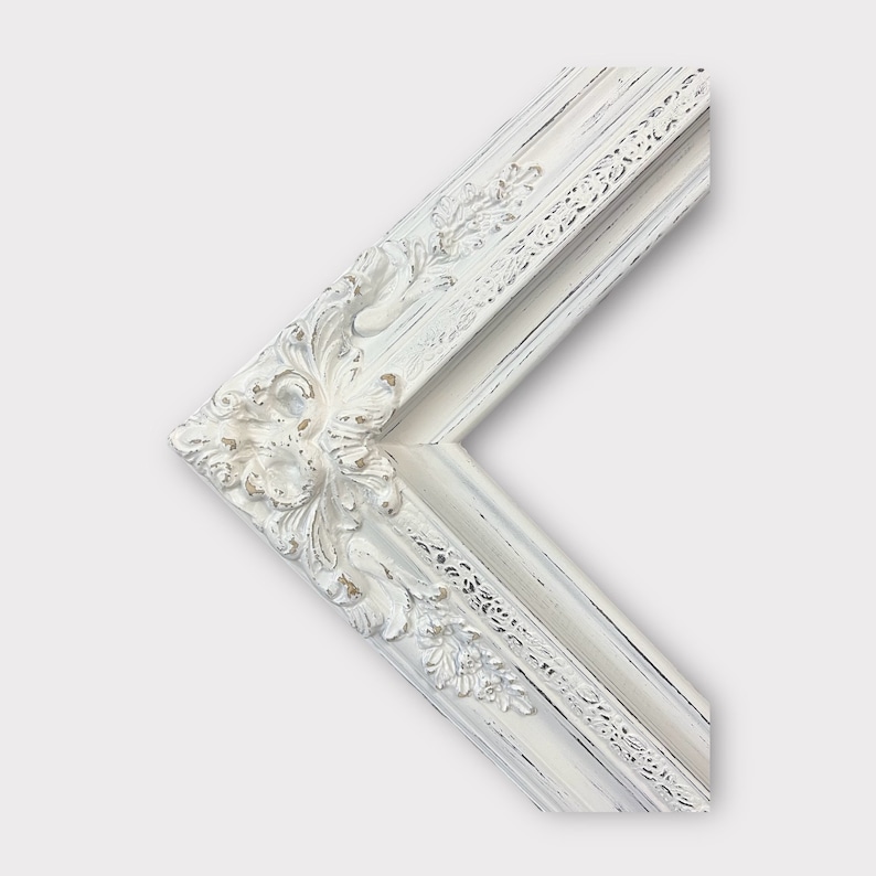 May include: Close-up of a corner of a white, ornate picture frame. The frame features detailed floral and scrollwork carvings with a distressed, antique finish. The frame's design is classic and elegant, suitable for various art styles.
