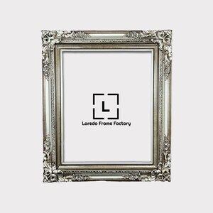May include: A silver ornate picture frame with a white mat. The frame has a decorative, ornate design with a raised edge. The frame is made of wood and has a distressed finish. The frame is empty and ready to be filled with a picture or artwork. The frame is made by Laredo Frame Factory.