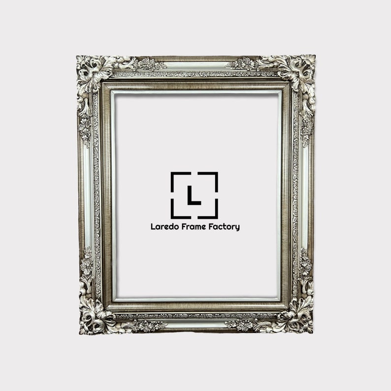 May include: A silver ornate picture frame with a white mat. The frame has a decorative, ornate design with a raised edge. The frame is made of wood and has a distressed finish. The frame is empty and ready to be filled with a picture or artwork. The frame is made by Laredo Frame Factory.