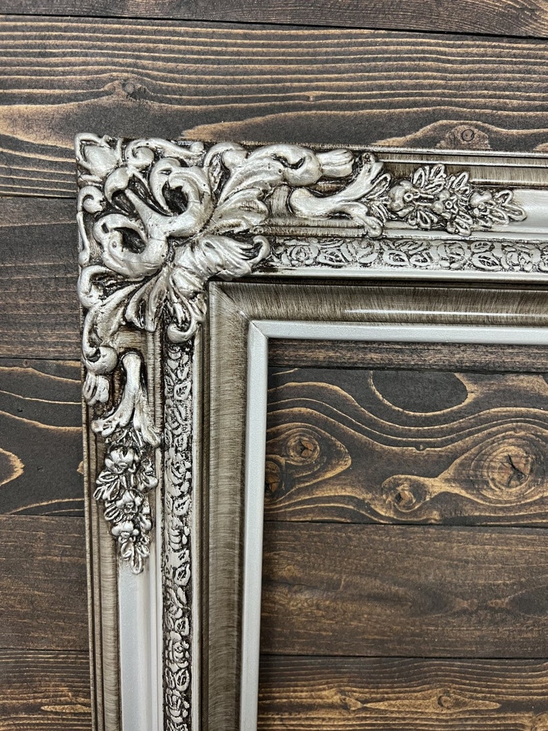 May include: A silver ornate picture frame with a detailed floral design. The frame is made of wood and has a distressed finish.