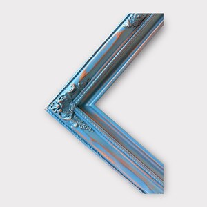 May include: A corner of a decorative picture frame with a distressed blue and copper finish. The frame has ornate detailing at the corner and a beveled edge. The frame is angled, showing the corner and a portion of the sides.