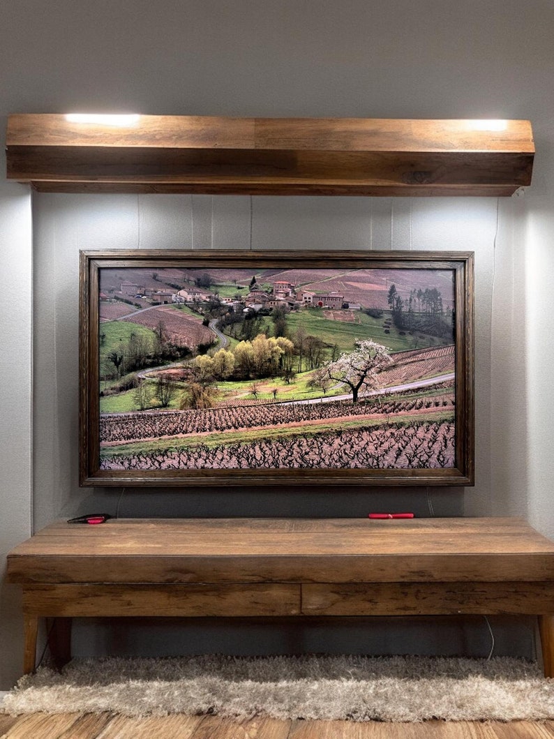 Puede incluir: Una fotograf&iacute;a de paisaje rectangular en un marco de madera oscura que representa un pueblo rural rodeado de colinas, vi&ntilde;edos y &aacute;rboles. La obra est&aacute; montada en una pared debajo de un estante horizontal de madera y sobre una mesa consola de madera.