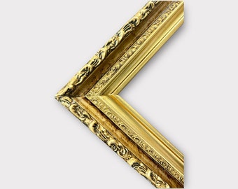 May include: A gold picture frame with ornate detailing. The frame is made of wood and has a gold finish. The frame is angled to show the corner.
