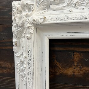 May include: A white, ornate picture frame with a distressed finish. The frame features detailed floral and scrollwork carvings at the corners and along the edges. The frame has a vintage, antique aesthetic.