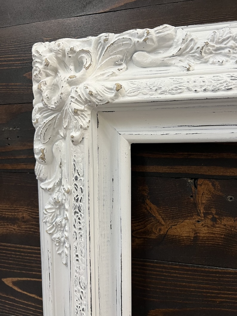 May include: A white, ornate picture frame with a distressed finish. The frame features detailed floral and scrollwork carvings at the corners and along the edges. The frame has a vintage, antique aesthetic.