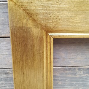 Peut inclure: Gros plan sur un cadre photo en bois peint en or avec une finition l&eacute;g&egrave;rement vieillie. Le cadre a un design simple et classique avec un bord sur&eacute;lev&eacute;.