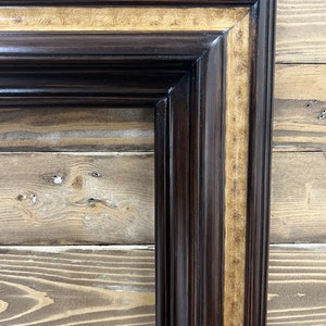 May include: A corner of a dark brown wooden picture frame with a glossy finish. The frame has a lighter, patterned wood inlay. The frame is rectangular with a detailed, layered design, showcasing a classic aesthetic.