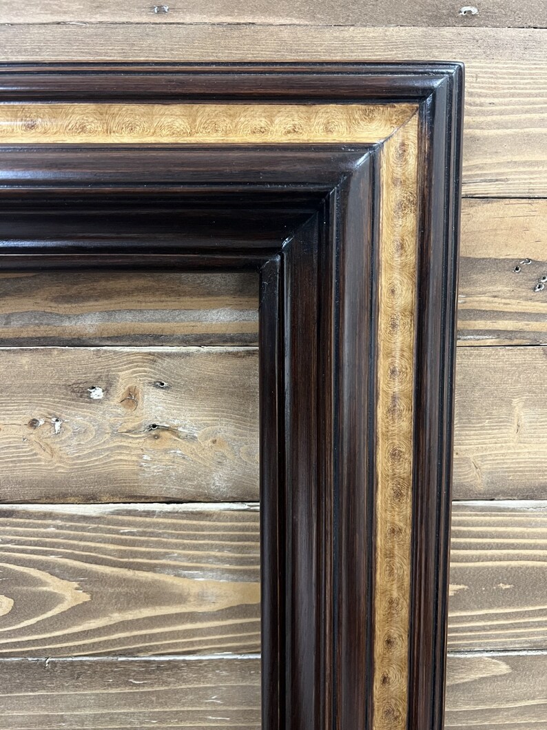 May include: A corner of a dark brown wooden picture frame with a glossy finish. The frame has a lighter, patterned wood inlay. The frame is rectangular with a detailed, layered design, showcasing a classic aesthetic.