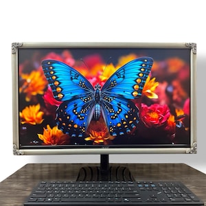 May include: A computer monitor with a silver ornate frame displaying a vibrant blue butterfly against a background of orange and red flowers. The monitor is positioned above a black computer keyboard on a dark wood-grain surface.
