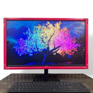 May include: A computer monitor featuring a bright pink, ornate, vintage-style frame surrounding the screen. The display shows a vibrant, multi-colored digital illustration of a tree with rainbow-colored leaves against a dark blue sky.