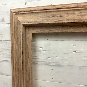 May include: A rectangular picture frame made of light brown wood with a visible grain texture and dark brown distressed detailing along the edges and grooves. The frame features a multi-tiered profile with clean mitered corners and a hollow center.