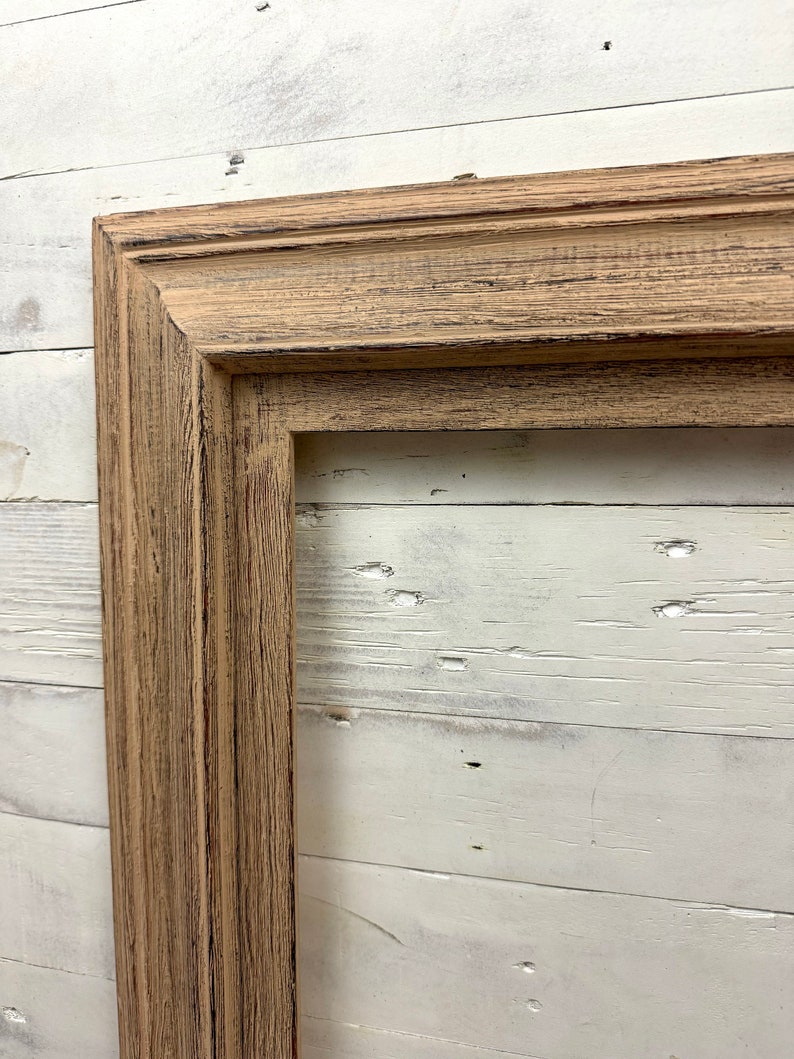 May include: A rectangular picture frame made of light brown wood with a visible grain texture and dark brown distressed detailing along the edges and grooves. The frame features a multi-tiered profile with clean mitered corners and a hollow center.