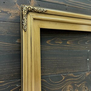 May include: A rectangular picture frame with a metallic gold finish and a smooth, multi-tiered profile. The top corner features an ornate, raised decorative accent with intricate scrollwork and leaf-like detailing, adding texture to the frame.