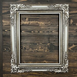 May include: A silver ornate picture frame with a decorative design. The frame is against a dark wood background.