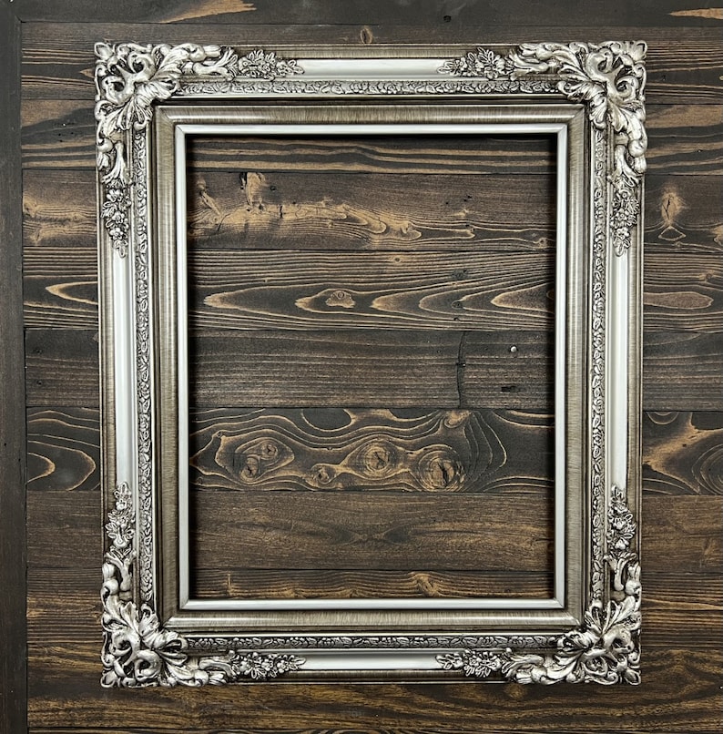 May include: A silver ornate picture frame with a decorative design. The frame is against a dark wood background.