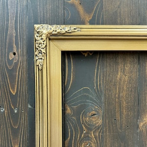 May include: A gold picture frame with ornate detailing on the top corner. The frame is against a dark wood background.
