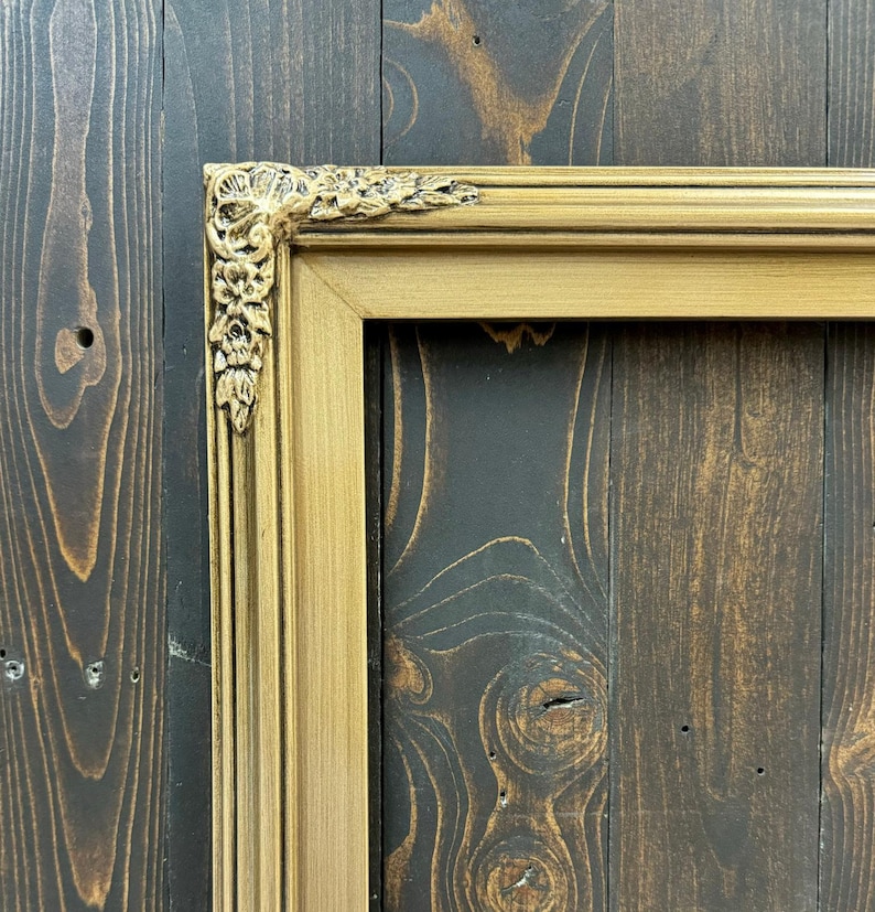 May include: A gold picture frame with ornate detailing on the top corner. The frame is against a dark wood background.