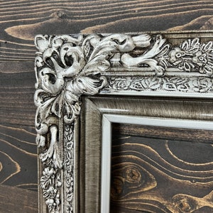 May include: A silver ornate picture frame with a detailed, intricate design. The frame has a distressed, aged look with a white inner border.