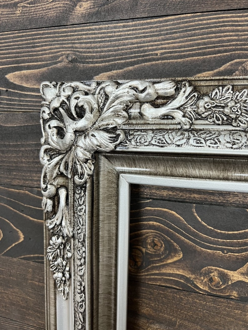 May include: A silver ornate picture frame with a detailed, intricate design. The frame has a distressed, aged look with a white inner border.