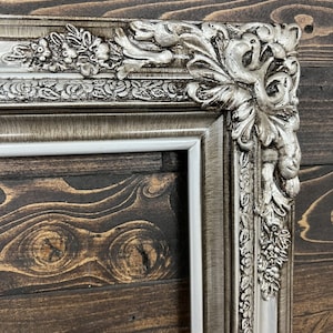 May include: A silver ornate picture frame with a white mat. The frame has a detailed design with floral and leaf motifs. The frame is against a dark wood background.