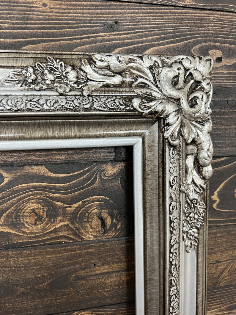 May include: A silver ornate picture frame with a white mat. The frame has a detailed design with floral and leaf motifs. The frame is against a dark wood background.
