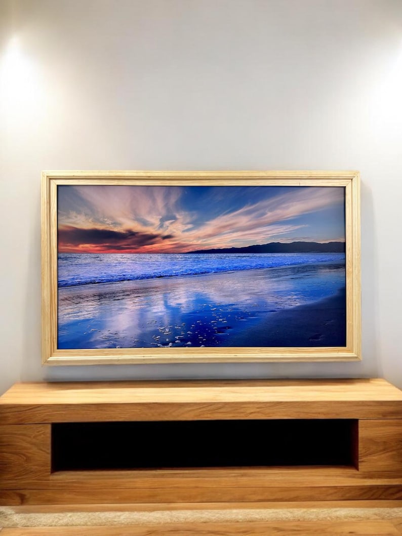 K&ouml;nnte beinhalten: Ein rechteckiger Fernsehbildschirm mit einem lebhaften Sonnenuntergang &uuml;ber dem Ozean, gerahmt in hellem Holz. Das Bild zeigt tiefblaue Wellen, ein dunkles Ufer und einen Himmel voller rosa, orangefarbener und violetter Wolkenstreifen bei D&auml;mmerung.