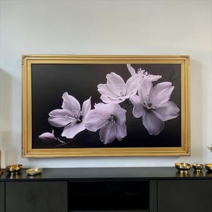 May include: A rectangular wall art piece featuring a cluster of pale lavender flowers against a solid black background, encased in a wide, ornate gold-toned frame. The floral arrangement includes several open blossoms and one closed bud.