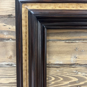 May include: Close-up of a corner of a dark brown wooden picture frame with a gold-colored inner border. The frame has a glossy finish and a classic, ornate design. The background is a light brown wooden surface.
