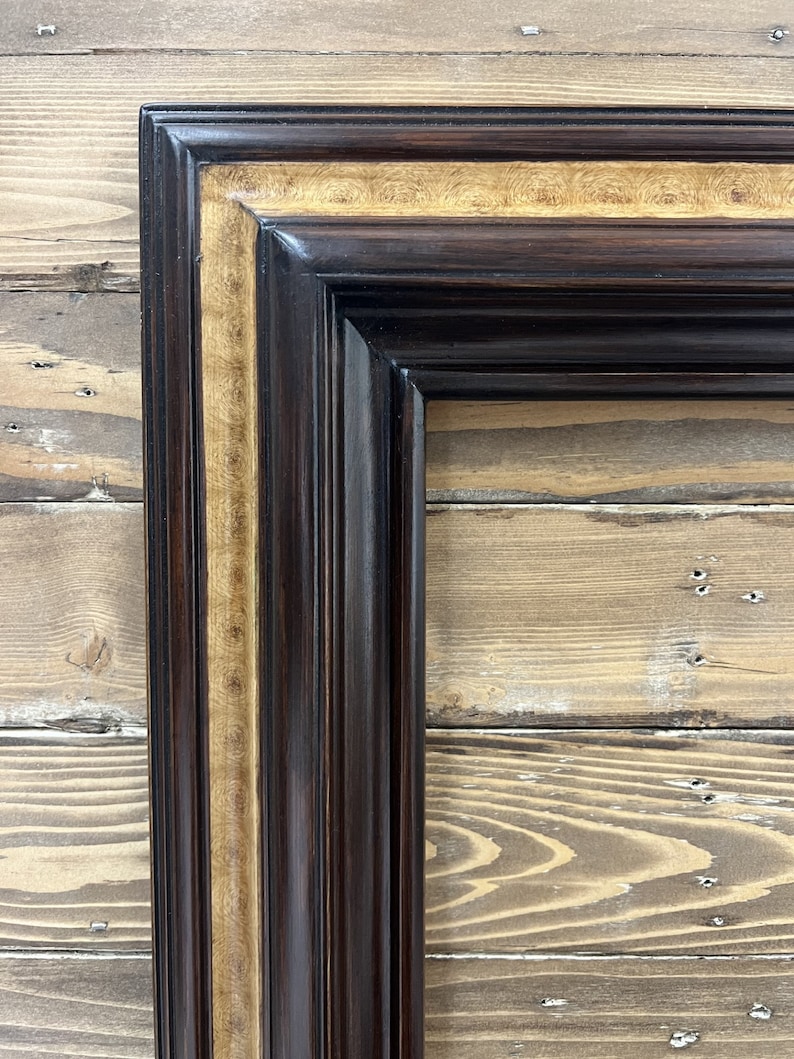 May include: Close-up of a corner of a dark brown wooden picture frame with a gold-colored inner border. The frame has a glossy finish and a classic, ornate design. The background is a light brown wooden surface.