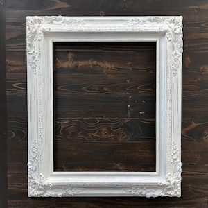 May include: A white, ornate picture frame with a distressed finish. The frame has detailed floral and scrollwork designs on the corners and edges. The frame is rectangular and empty, ready for a photo or artwork.