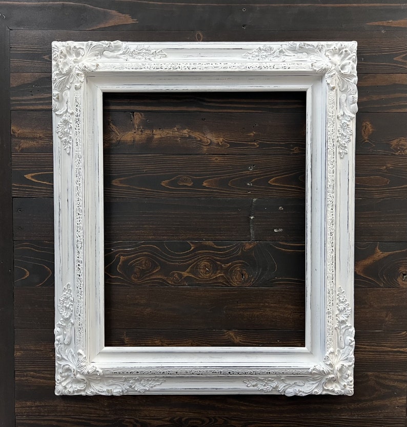 May include: A white, ornate picture frame with a distressed finish. The frame has detailed floral and scrollwork designs on the corners and edges. The frame is rectangular and empty, ready for a photo or artwork.