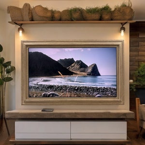 May include: A television mounted on a wall with a wide, textured, light-colored wooden frame. Above the television is a wooden shelf holding several woven baskets filled with greenery. Below the television is a long, low console with white drawers.