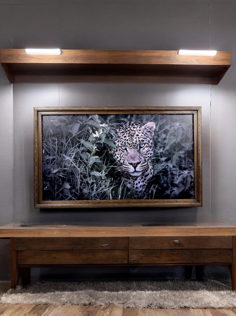 Puede incluir: Una pieza de arte mural rectangular enmarcada que presenta una fotograf&iacute;a en blanco y negro de un leopardo mirando a trav&eacute;s de un follaje denso. La obra est&aacute; en un marco de madera marr&oacute;n oscuro y montada en una pared gris bajo un estante.