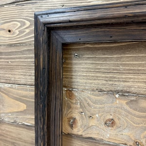 Puede incluir: Un marco rectangular fabricado en madera te&ntilde;ida de oscuro con un patr&oacute;n de veta visible. El marco presenta una uni&oacute;n en inglete y una superficie texturizada con peque&ntilde;as hendiduras, mostrando un acabado marr&oacute;n profundo en toda su estructura.