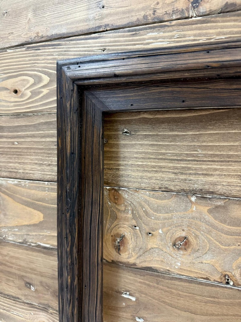 Puede incluir: Un marco rectangular fabricado en madera te&ntilde;ida de oscuro con un patr&oacute;n de veta visible. El marco presenta una uni&oacute;n en inglete y una superficie texturizada con peque&ntilde;as hendiduras, mostrando un acabado marr&oacute;n profundo en toda su estructura.