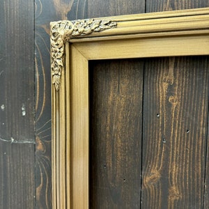 May include: A gold-colored picture frame with ornate detailing on the corners. The frame is against a dark wood background.
