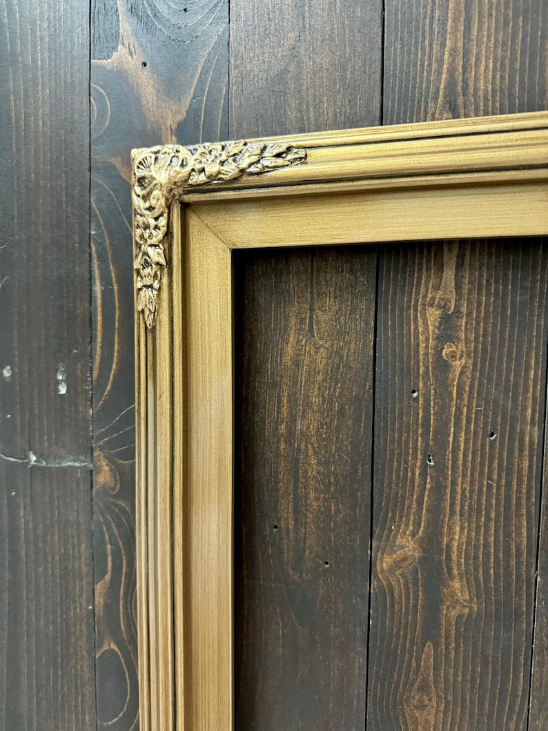 May include: A gold-colored picture frame with ornate detailing on the corners. The frame is against a dark wood background.